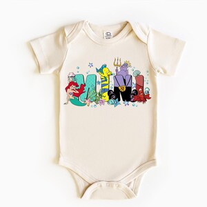 Puede incluir: Body de beb&eacute; color crema con la palabra "MAMI" en letras coloridas de estilo caricatura. Las letras est&aacute;n decoradas con personajes de una popular pel&iacute;cula animada, incluyendo una sirena, un pez y criaturas marinas.