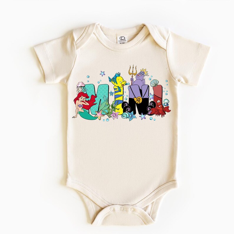Puede incluir: Body de beb&eacute; color crema con la palabra "MAMI" en letras coloridas de estilo caricatura. Las letras est&aacute;n decoradas con personajes de una popular pel&iacute;cula animada, incluyendo una sirena, un pez y criaturas marinas.