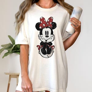 Puede incluir: Camiseta color crema con un gr&aacute;fico desgastado de Minnie Mouse. El personaje de dibujos animados est&aacute; en negro, blanco y rojo, con su ic&oacute;nico lazo de lunares. La camiseta tiene un corte holgado y mangas cortas, ideal para el uso diario.