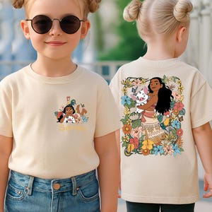 May include: Beige t-shirt featuring a colorful graphic of a cartoon character with long black hair, holding a pig, surrounded by flowers. The front of the shirt has a similar design with the name "Serena" in gold lettering. The shirt is worn by a young girl.