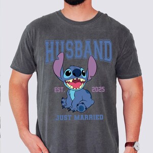 Puede incluir: Camiseta gris oscuro con un personaje de dibujos animados azul y la palabra "HUSBAND" en letras azules. La camiseta tambi&eacute;n presenta el texto "EST. 2025" y "JUST MARRIED" debajo del personaje.