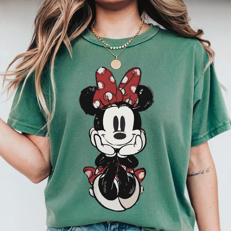 Puede incluir: Camiseta verde con un gr&aacute;fico de Minnie Mouse. El gr&aacute;fico muestra a Minnie Mouse con un lazo rojo de lunares y un dise&ntilde;o en blanco y negro. La camiseta tiene cuello redondo y mangas cortas.