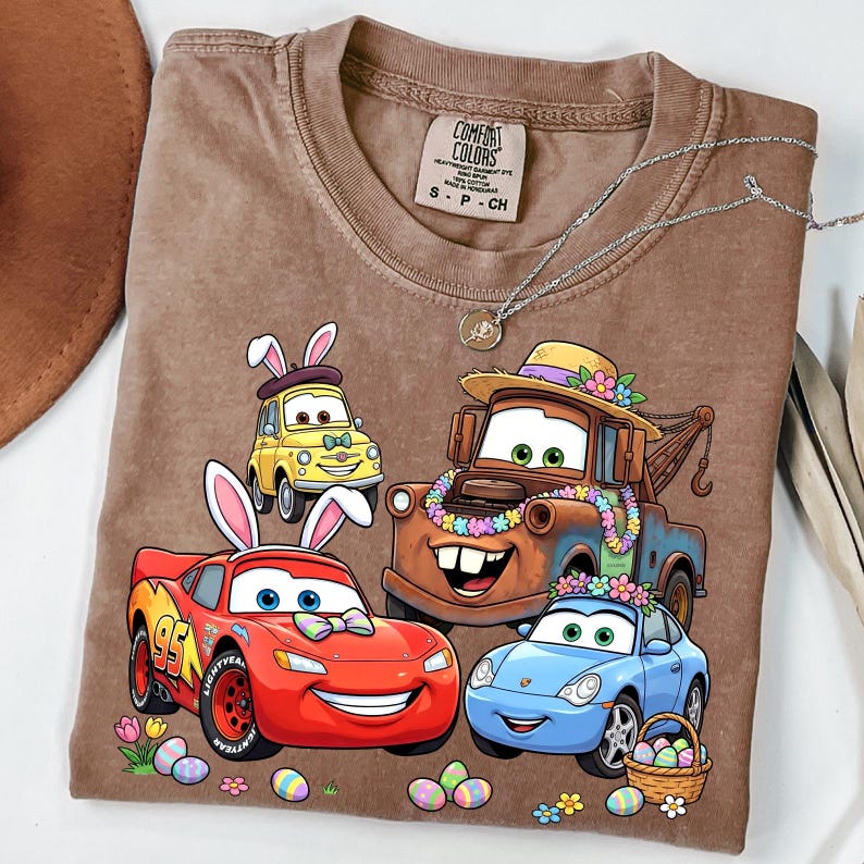 May include: Brown t-shirt featuring cartoon cars with bunny ears and Easter eggs. The shirt has the text "Comfort Colors" on the tag. A silver necklace is draped across the shirt.
