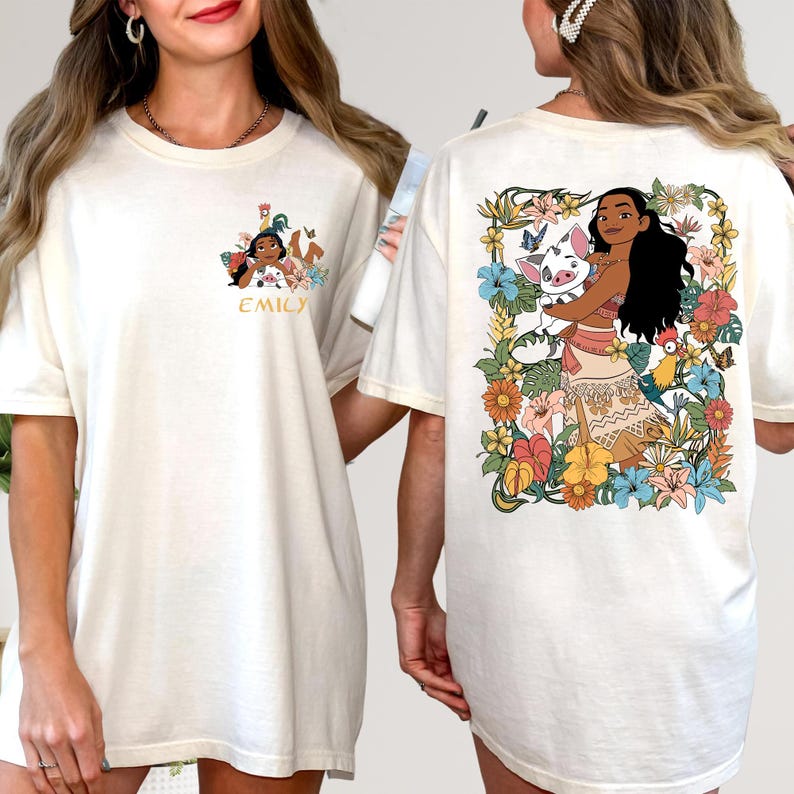 May include: Cream-colored t-shirts with colorful graphic designs. One shirt features a design with the name "EMILY" and a cartoon character. The other shirt has a floral design with a cartoon character and a pig.