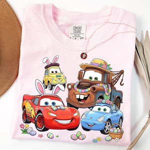 May include: Pink t-shirt featuring cartoon cars decorated with Easter bunny ears and floral garlands. The design includes Lightning McQueen, Mater, and other characters, along with Easter eggs and flowers. The shirt has the Comfort Colors brand tag.