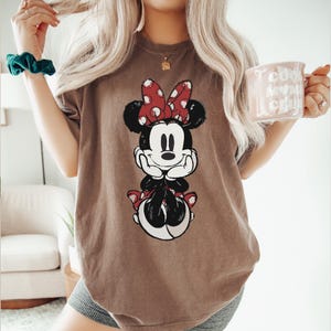 Puede incluir: Camiseta marr&oacute;n con un gr&aacute;fico de Minnie Mouse. El personaje de dibujos animados tiene un lazo rojo con lunares y est&aacute; sentado. La camiseta es de color neutro y tiene un corte holgado. La imagen tambi&eacute;n muestra una taza con texto.