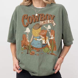 May include: A sage green t-shirt featuring a graphic print of two figures in cowboy hats sitting in a desert landscape with cacti and a horse. The text reads "COWBOY LIKE ME" at the top and "I KNEW YOU" in a circular pattern around the figures.