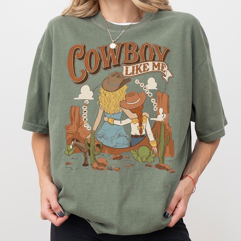 May include: A sage green t-shirt featuring a graphic print of two figures in cowboy hats sitting in a desert landscape with cacti and a horse. The text reads "COWBOY LIKE ME" at the top and "I KNEW YOU" in a circular pattern around the figures.