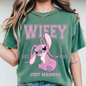 Puede incluir: Camiseta verde con la palabra "Wifey" en letras rosas en bloque sobre un personaje de dibujos animados rosa. Debajo del personaje, las palabras "Just Married" est&aacute;n impresas en rosa. El a&ntilde;o "2025" est&aacute; impreso a la derecha del personaje.