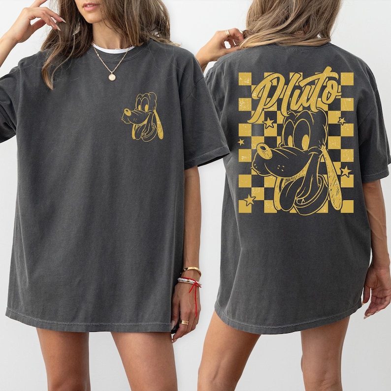 Puede incluir: Camiseta gris oscuro con un dise&ntilde;o dorado de Pluto. La parte delantera tiene una peque&ntilde;a imagen de Pluto, mientras que la parte trasera muestra un dise&ntilde;o m&aacute;s grande de Pluto con la palabra "Pluto" y un fondo a cuadros.