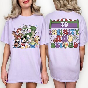May include: A light purple t-shirt featuring a colorful graphic of Toy Story characters on the front. The back of the shirt has the text "To Infinity and Beyond" in a retro font, with a rocket ship and other graphics.