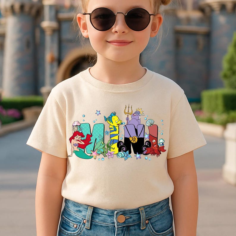 Puede incluir: Una camiseta beige con la palabra "MIMI" en letras coloridas de estilo caricatura. Las letras est&aacute;n decoradas con personajes de una popular pel&iacute;cula de animaci&oacute;n. La camiseta la lleva una persona joven.