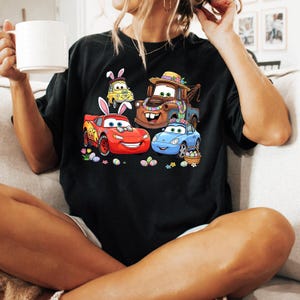 May include: Black t-shirt featuring a colorful graphic of animated cars decorated for Easter. The design includes a red race car, a blue car, a yellow car, and a brown tow truck, all with Easter-themed accessories and surrounded by Easter eggs.
