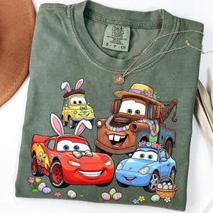May include: Green t-shirt featuring cartoon cars with bunny ears and Easter eggs. The design includes a red car with the number 95, a yellow car, a brown tow truck, and a blue car. The shirt has a Comfort Colors tag.