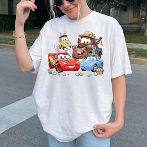 May include: White t-shirt featuring a colorful graphic of animated cars decorated for Easter. The design includes a red car, a blue car, a brown tow truck, and a yellow car, all with Easter-themed decorations and surrounded by Easter eggs.