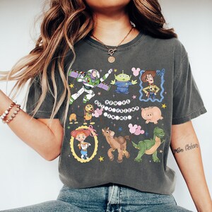 May include: Dark gray t-shirt featuring colorful cartoon characters from a popular animated movie. The shirt includes Buzz Lightyear, an alien, Woody, Jessie, a dog, a pig, a dinosaur, and a horse. The text "You've Got a Friend in Me" is also printed on the shirt.