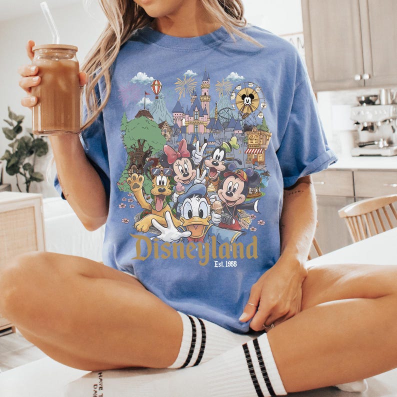 Puede incluir: Una camiseta azul de manga corta con un estampado gr&aacute;fico colorido de varios personajes de dibujos animados, incluyendo a Mickey Mouse, Minnie Mouse, Donald Duck, Goofy y Pluto frente a un castillo, con el texto Disneyland Est. 1955 impreso debajo.