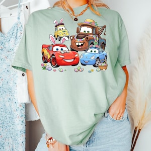 May include: A light green t-shirt featuring a colorful graphic of animated cars decorated for Easter. The cars include a red race car, a blue car, a yellow car, and a brown tow truck, all with Easter-themed decorations.