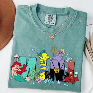 Puede incluir: Una camiseta Comfort Colors verde mar con la palabra "MOM" y personajes de La Sirenita. Se representan a Ariel, Flounder, &Uacute;rsula y Sebasti&aacute;n. Un collar plateado con un colgante est&aacute; sobre la camiseta.