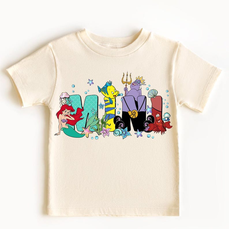 Puede incluir: Camiseta color crema con la palabra "MAMI" en letras coloridas de estilo caricatura. Cada letra est&aacute; decorada con personajes de una popular pel&iacute;cula animada, incluyendo una sirena, un pez amarillo y una bruja marina p&uacute;rpura.