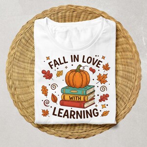 May include: White t-shirt with a fall-themed graphic. The design features a stack of books topped with a pumpkin, surrounded by autumn leaves and the text "FALL IN LOVE WITH LEARNING". The shirt is displayed on a woven, circular surface.