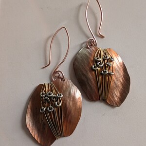 May include: A pair of handcrafted copper earrings. Each earring features a leaf-shaped design with a textured surface and a cluster of small, silver-colored rings and gold-colored wire accents. The ear wires are copper.