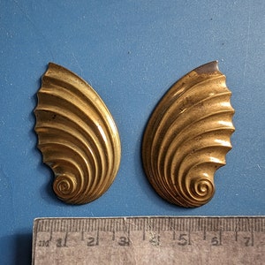May include: Two gold-colored metal shell-shaped decorative pieces. Each piece has a spiral design in the center and a textured surface.