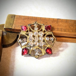 May include: A vintage gold-toned brooch with a starburst design. The brooch features clear and red rhinestones set in a filigree pattern. The brooch is approximately 2 inches in diameter.