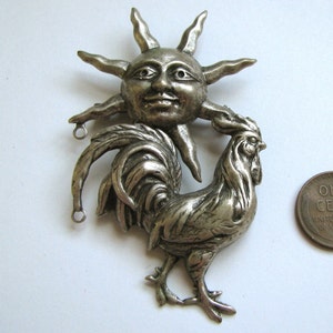 May include: Silver tone pendant featuring a rooster with a sun face above it. The rooster is standing on one leg and has its tail feathers spread out. The sun has a smiling face with eyes and a nose.