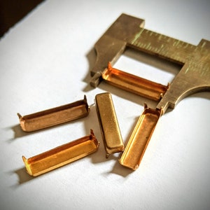 May include: Five small metal pieces, including four rectangular pieces with a copper finish and one rectangular piece with a gold finish. The pieces are arranged on a white surface.