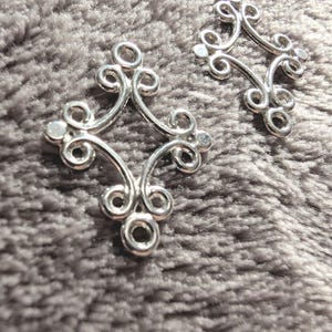 May include: Two silver metal decorative connectors with a swirling design. The connectors are shaped like a diamond with rounded corners.