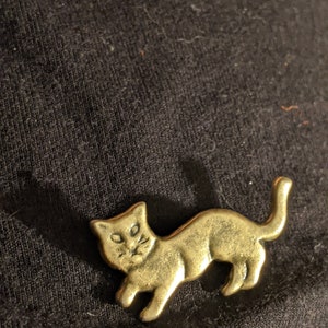 May include: A gold-colored metal pin shaped like a cat. The cat is in a running pose with its tail raised.