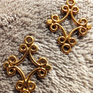 May include: Two gold-colored metal decorative pieces with a diamond shape and intricate swirls. The pieces are on a fuzzy gray surface.
