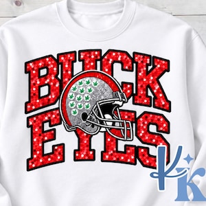 May include: A white crewneck sweatshirt featuring the text BUCKEYES in a red, sequin-style pattern. Centered is a silver football helmet graphic decorated with small green leaf icons. A blue logo with the letters KK is visible in the bottom right.