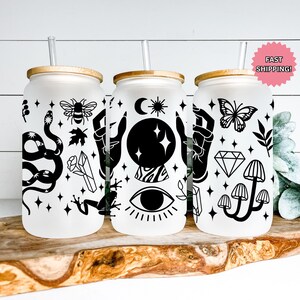 May include: Three frosted glass tumblers with bamboo lids and straws. Each tumbler features black and white mystical designs, including a snake, bee, crystal, frog, hands, crystal ball, eye, butterfly, diamond, and mushrooms. The tumblers sit on a wooden surface.