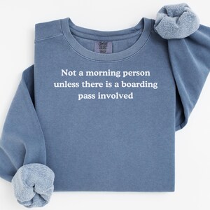 May include: A blue crew neck sweatshirt with the white text: "Not a morning person unless there is a boarding pass involved". The sleeves are cuffed, and a matching scrunchie is on the shoulder. The sweatshirt is a Comfort Colors brand.
