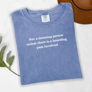 May include: A blue t-shirt with the text "Not a morning person unless there is a boarding pass involved" in white. The shirt is folded and has a Comfort Colors tag. A brown hat is partially visible in the background.