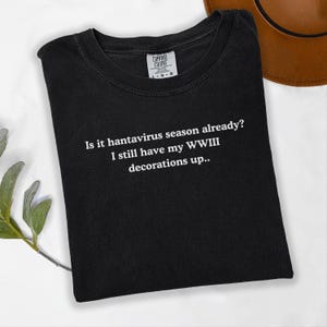 May include: Black t-shirt with the text "Is it hantavirus season already? I still have my WWIII decorations up.." in white. The shirt has a crew neck and a Comfort Colors tag. A brown hat is partially visible in the background.