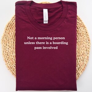 May include: Burgundy t-shirt with the text "Not a morning person unless there is a boarding pass involved" in white. The shirt is folded and placed on a woven, circular placemat. A white tag is visible on the shirt.