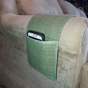 May include: A green fabric couch armrest organizer with a pocket holding a white and black smartphone. The organizer is designed to fit over the arm of a beige couch, providing storage for a phone or other small items.