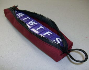 May include: A burgundy zippered pouch with a black zipper and a black pull tab. The pouch is open and contains a purple pill organizer with the days of the week labeled in white.