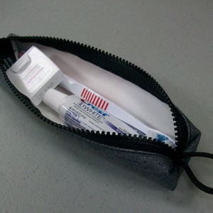 May include: A denim-colored zippered pouch containing dental hygiene products. Inside, a white toothbrush with red bristles, a tube of Crest 3D White toothpaste, and a box of Listerine dental floss are visible. The pouch has a black zipper and a black loop.