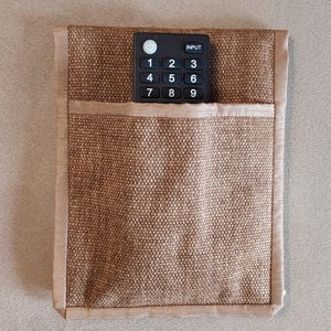 Könnte beinhalten: Ein brauner, gewebter Fernbedienungshalter mit einer Tasche und einer schwarzen Fernbedienung darin. Die Fernbedienung hat weiße Zahlen und eine "INPUT"-Taste. Der Halter hat eine hellbraune Umrandung.