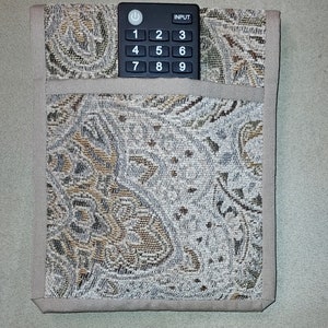 May include: A beige fabric remote control holder with a black remote control inside. The remote control has a power button, input button, and number keys 1 through 9.