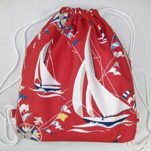 May include: Red drawstring backpack with a nautical theme. The bag features white sailboats, flags, and a compass rose design. White drawstrings and a red fabric loop complete the design.