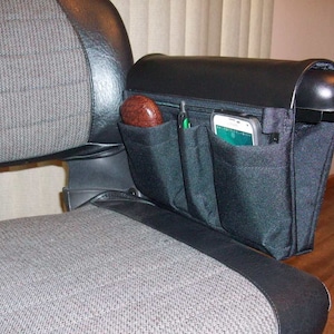 Puede incluir: Organizador de reposabrazos negro con múltiples bolsillos para silla de ruedas o scooter. El organizador tiene un bolsillo para teléfono móvil, un bolsillo para bolígrafos y un bolsillo más grande para otros artículos.