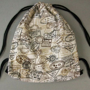 May include: A white drawstring bag with black straps. The bag is covered in brown and black stamp-like designs with text such as "MALI", "MEXICO", "BRAZIL", and "NARITA".