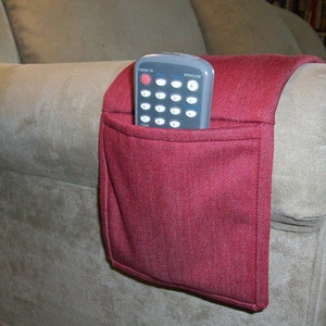 May include: A burgundy fabric remote control holder attached to the arm of a beige couch. The holder has a pocket to hold a remote control. A black remote control is in the pocket.