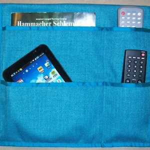 May include: A teal blue fabric organizer with three pockets. The top pocket has a magazine with the text "Hammacher Schlem" visible. The middle pocket has a tablet computer. The bottom pocket has a black remote control.