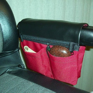 May include: A red zippered pouch with two pockets attached to a black armrest. The pouch has a black zipper and a black leather flap. The pouch is filled with a white wallet and a brown case.